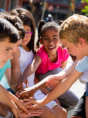 Children gather smiling with their hands in the middle on top of one another, as a team.